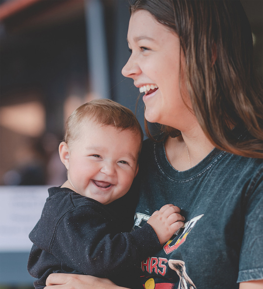 A child smiling at the camera whilst hugging its mum