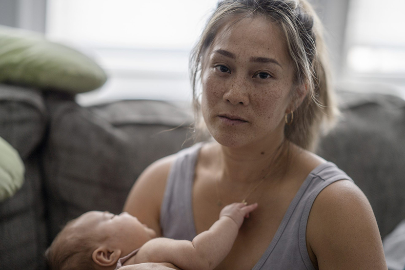 A middle aged mother sitting on her couch holds her baby in her arm with a tired and stressed expression.